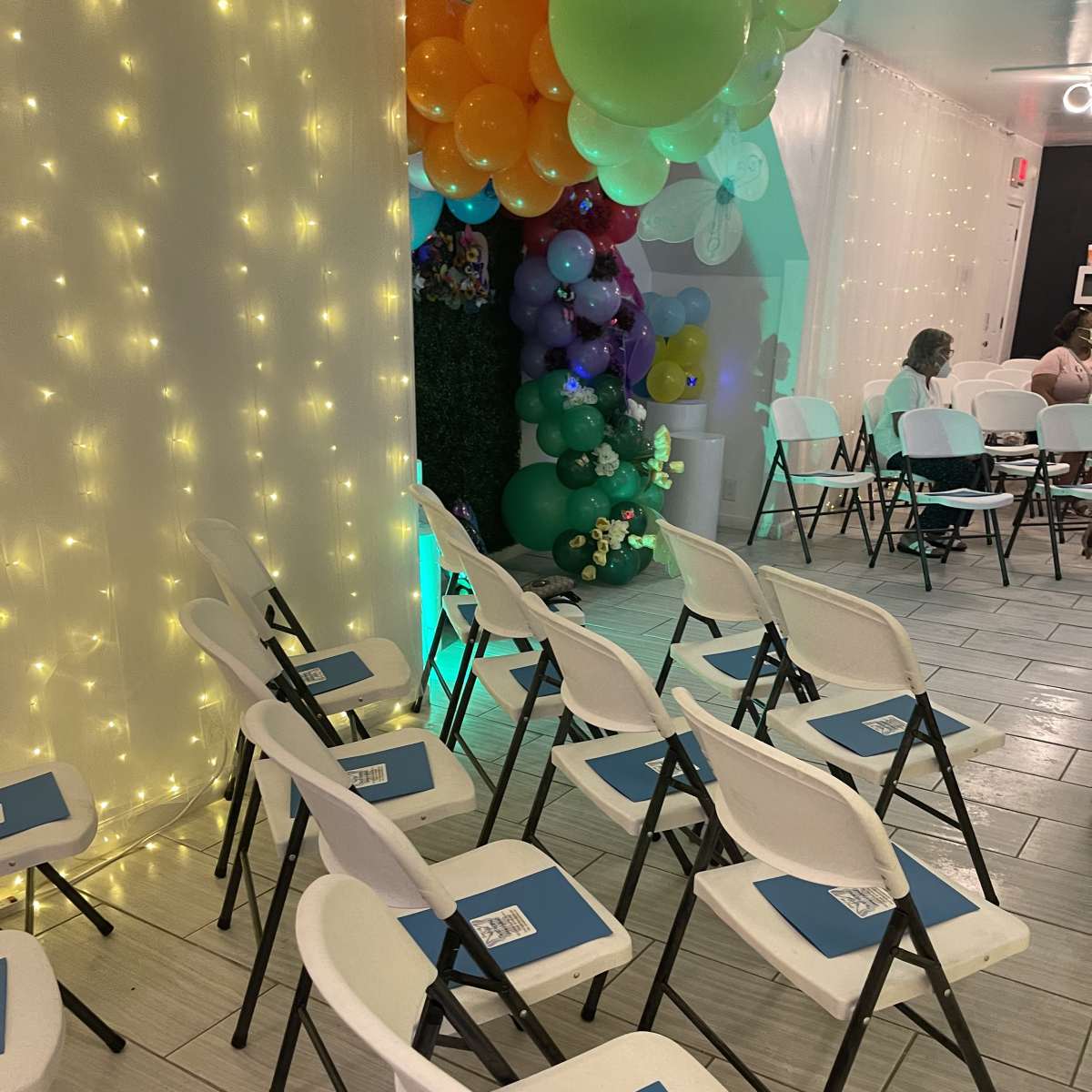 A row of white folding chairs with blue folders on them, arranged near a colorful balloon decoration in a brightly lit room.