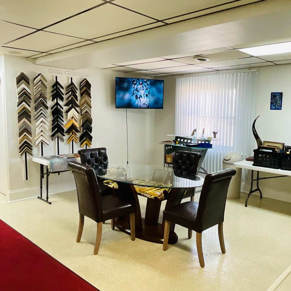 A room with a glass table and two chairs, a wall of picture frames, a television, and tables displaying framed artwork and other items.