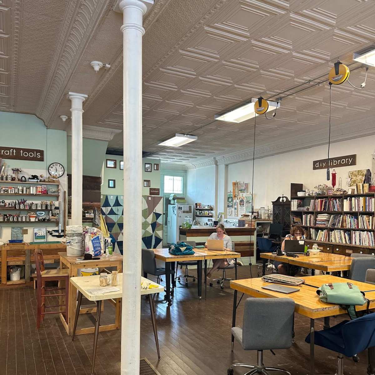 A spacious craft studio with several work tables, shelves filled with tools and books, and individuals engaged in various activities.
