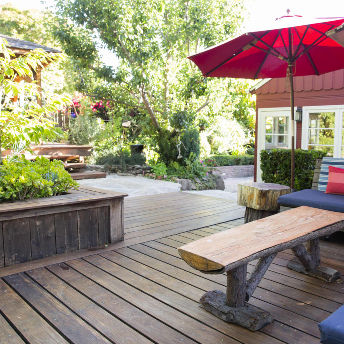 A wooden deck features a seating area with a blue couch and a rustic table beneath a red umbrella, surrounded by greenery and a red building.