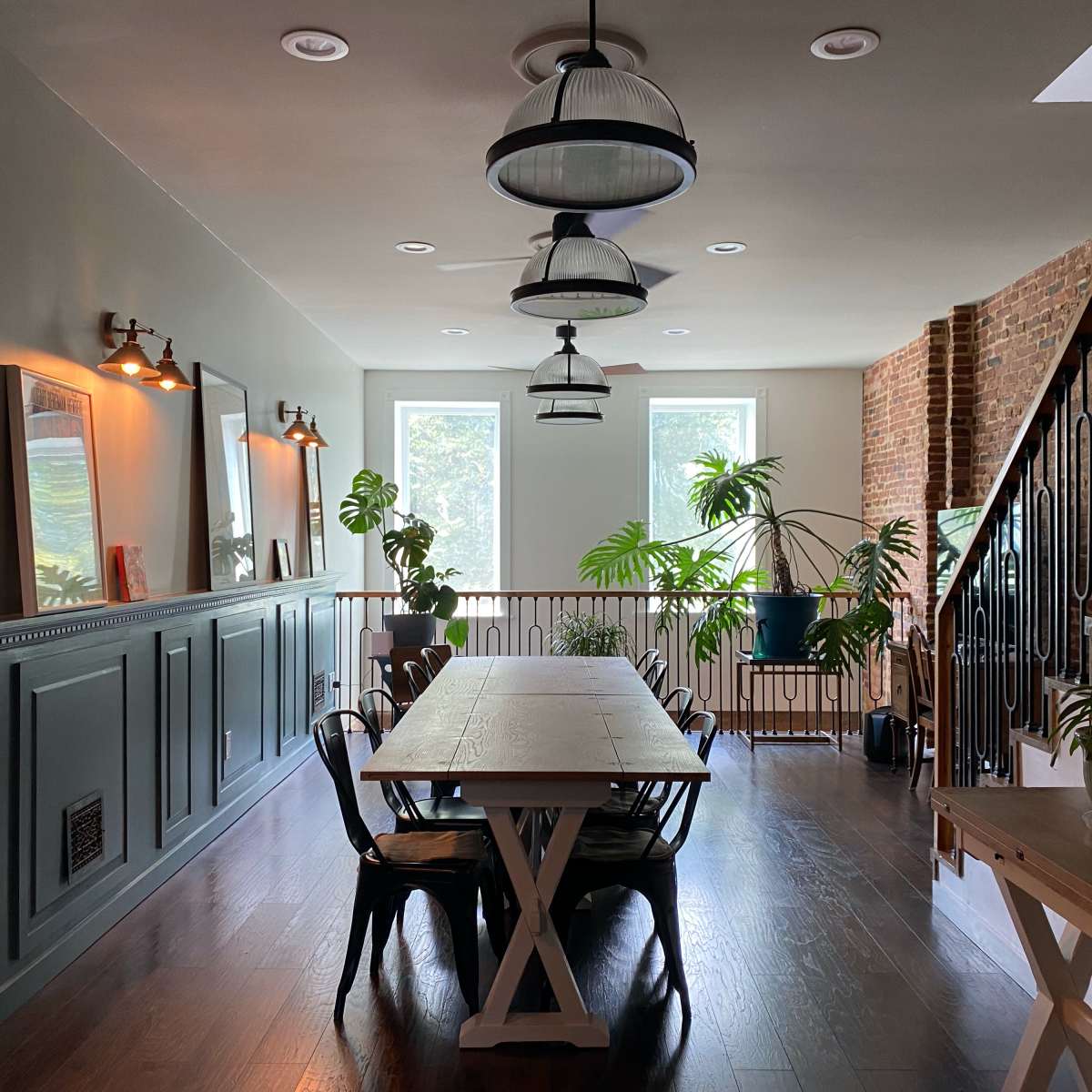 A dining area features a long wooden table surrounded by black chairs, with large windows allowing natural light and indoor plants decorating the space.