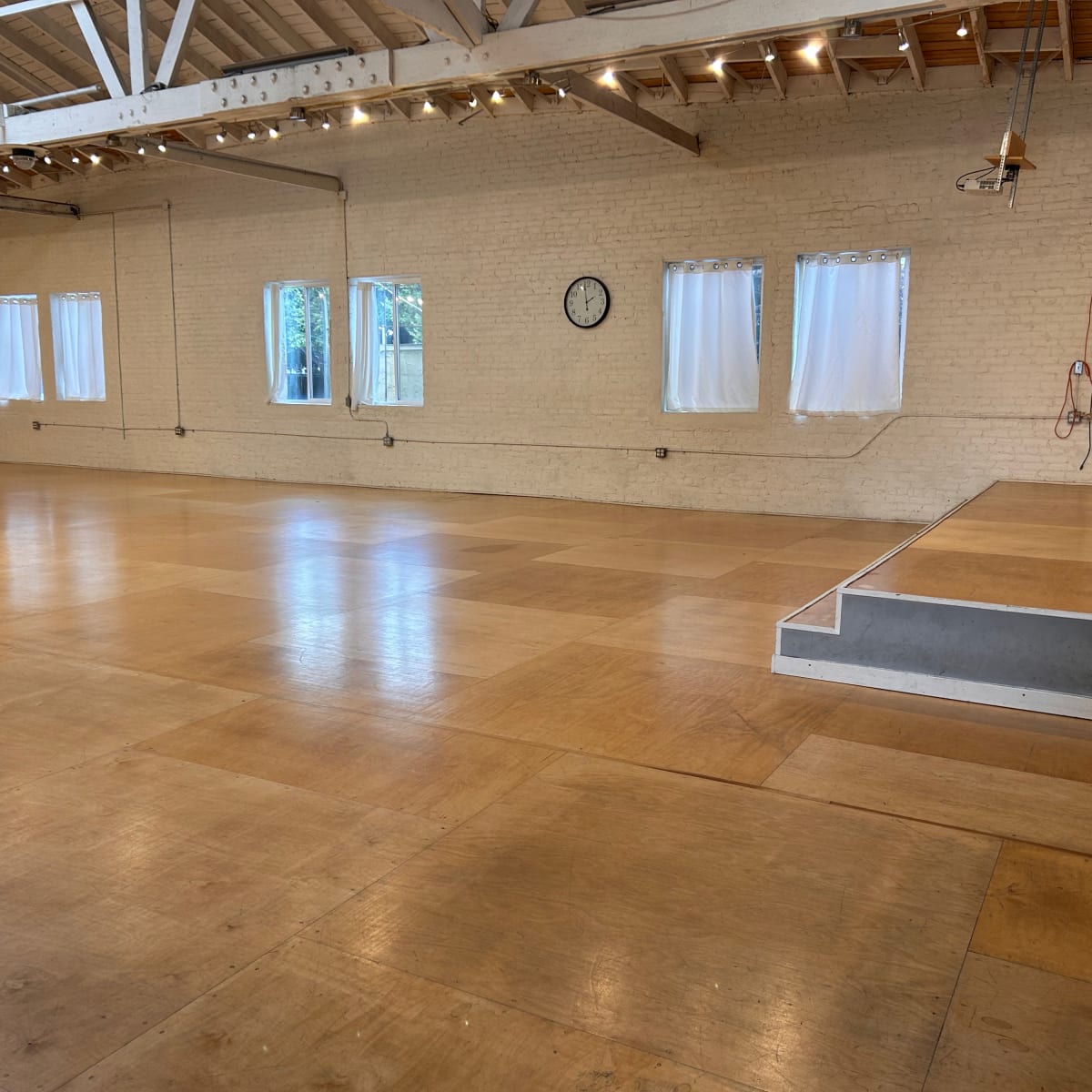 A spacious, empty interior room with a wooden dance floor, large windows, and a clock on the wall.