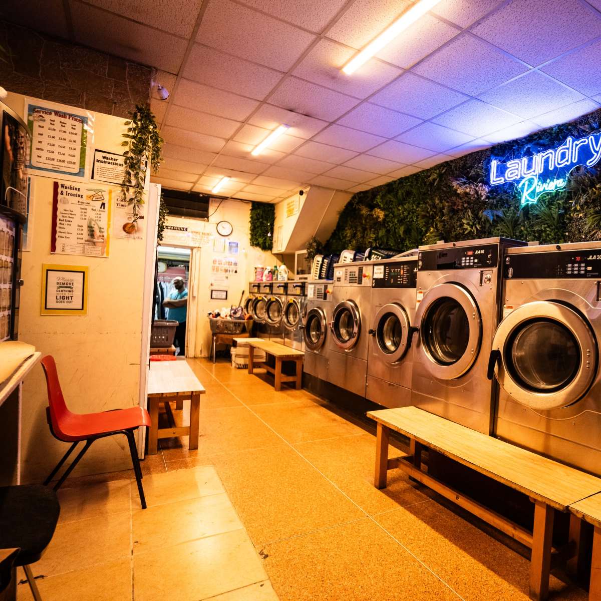 A row of washing machines in a laundromat, with neon signage and greenery decor overhead.
