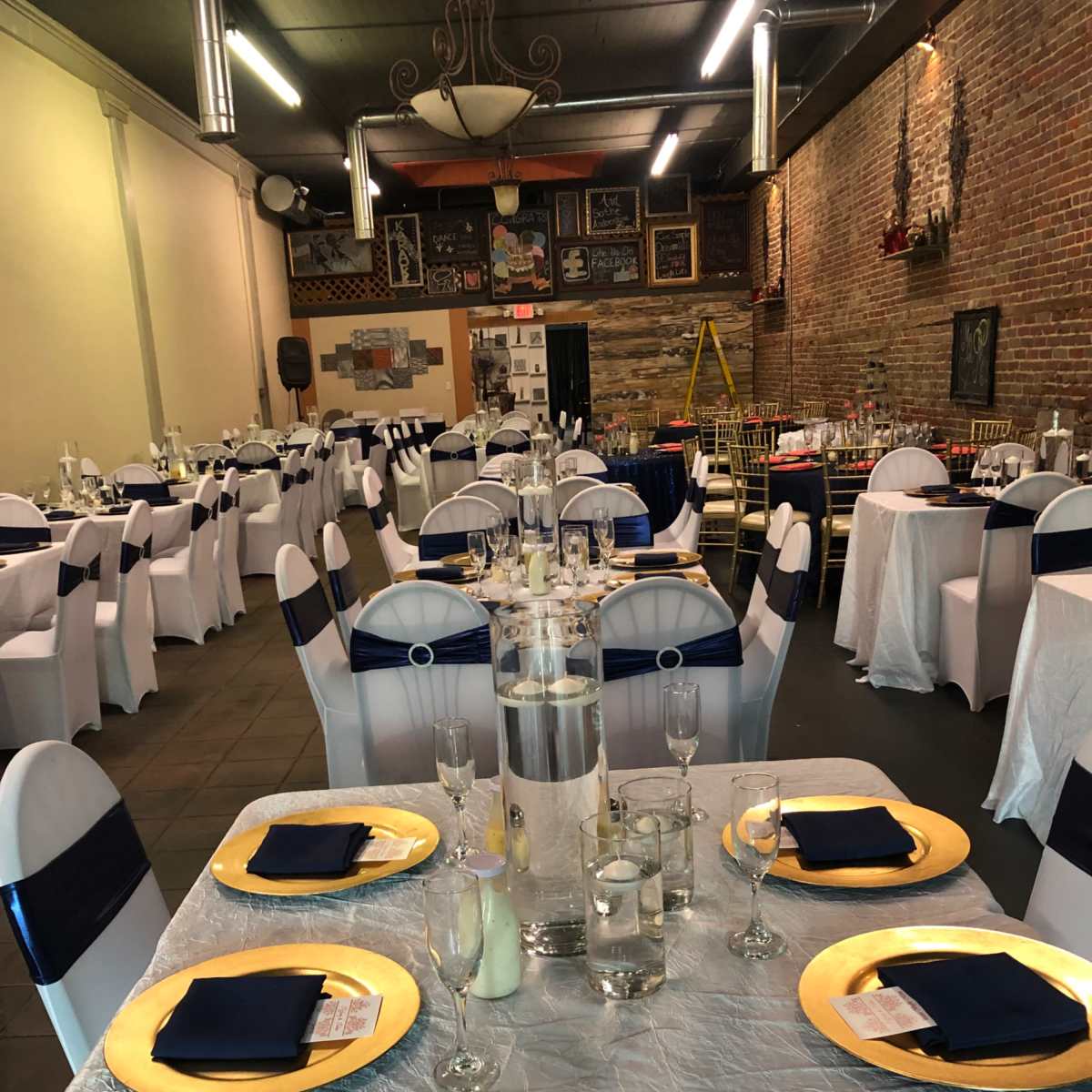 A banquet hall set for an event, featuring elegantly arranged tables with white tablecloths, gold chargers, and navy blue napkins, along with a brick wall and decorative elements in the background.
