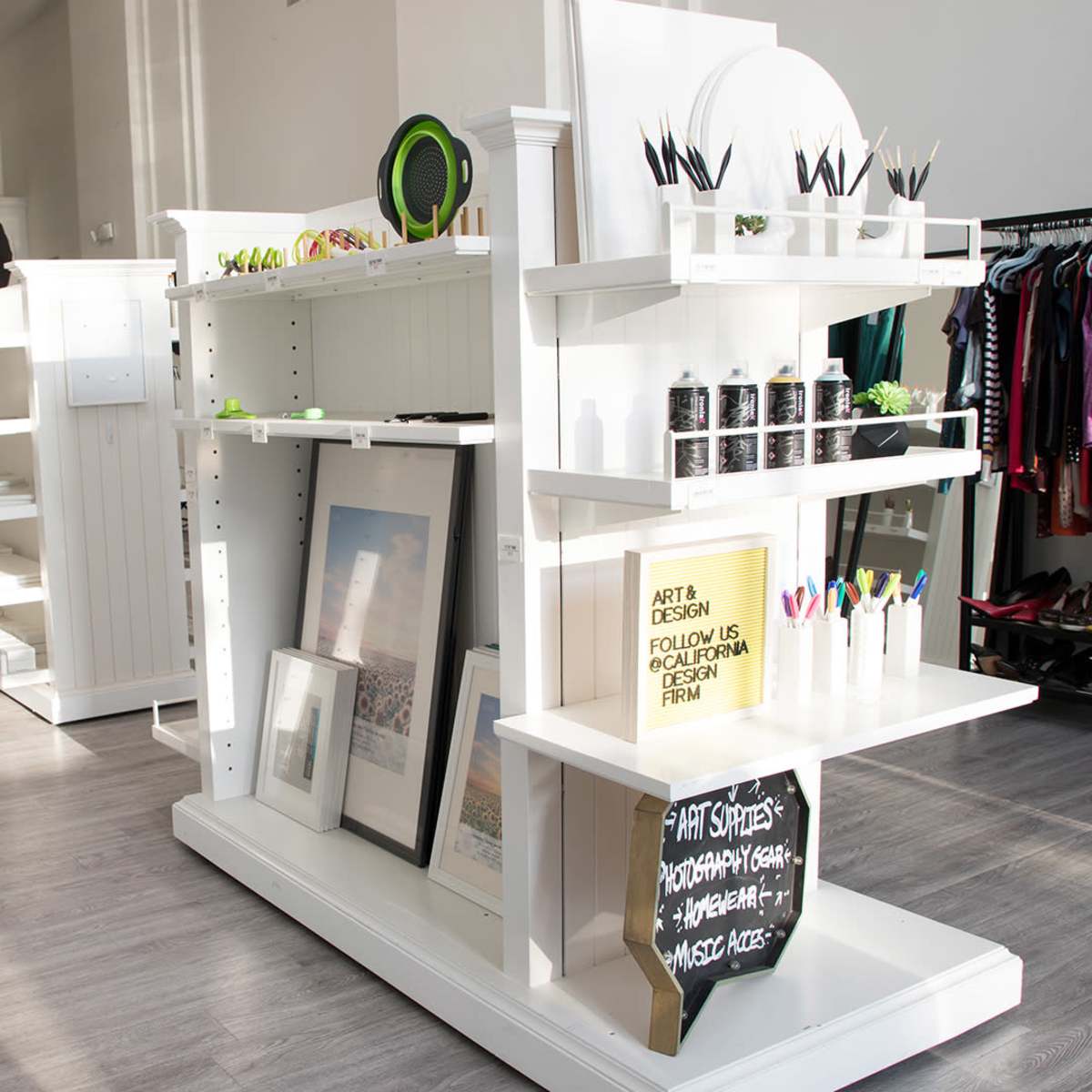 A white interior space featuring display shelves with art supplies and framed art, alongside a clothing rack and a photography setup in the background.