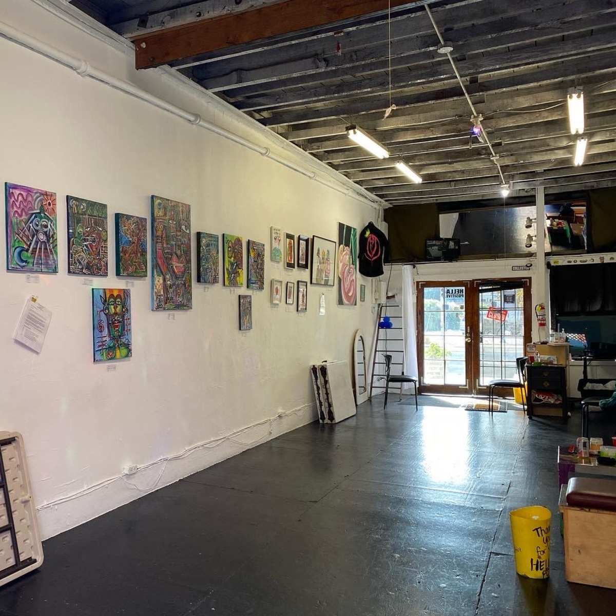 An art gallery featuring colorful paintings displayed on a white wall, with a folding table and a ladder visible in the interior space.