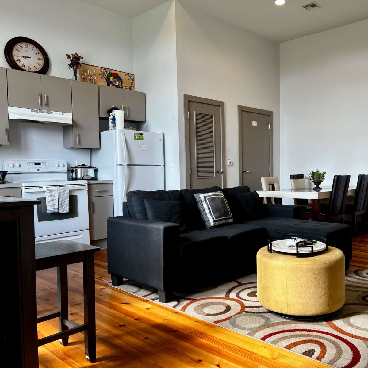 A modern living space with a black sofa, a round yellow ottoman, a dining table with chairs, and a kitchen area in the background.