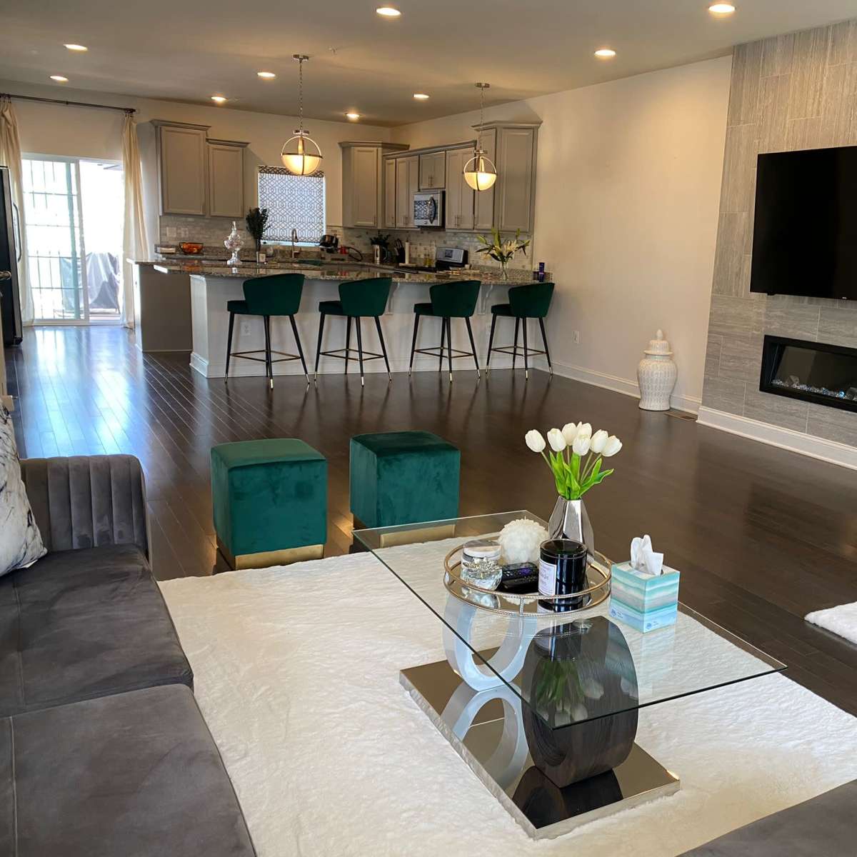 A modern living room with a glass coffee table and a sectional sofa, leading to an open-concept kitchen with bar seating.