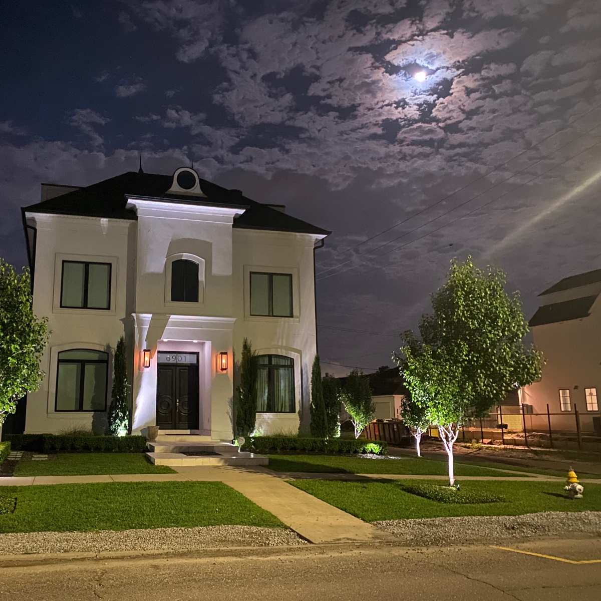 A well-lit, two-story white house stands at night under a partly cloudy sky, with two small trees illuminated on either side of the pathway leading to the front door.