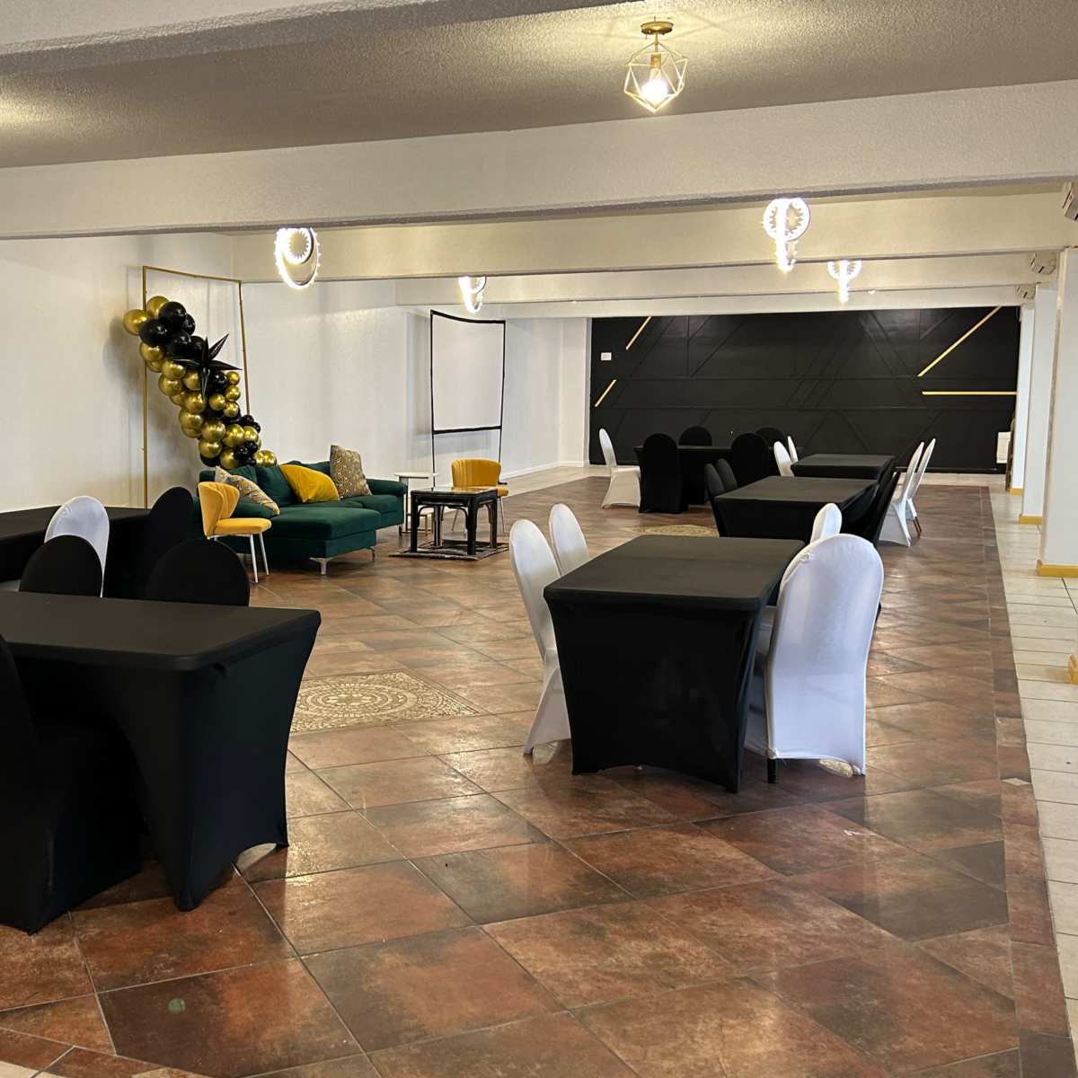 A spacious event room with several tables covered in black and white tablecloths, along with a seating area featuring a green couch and decorative balloons on one side.