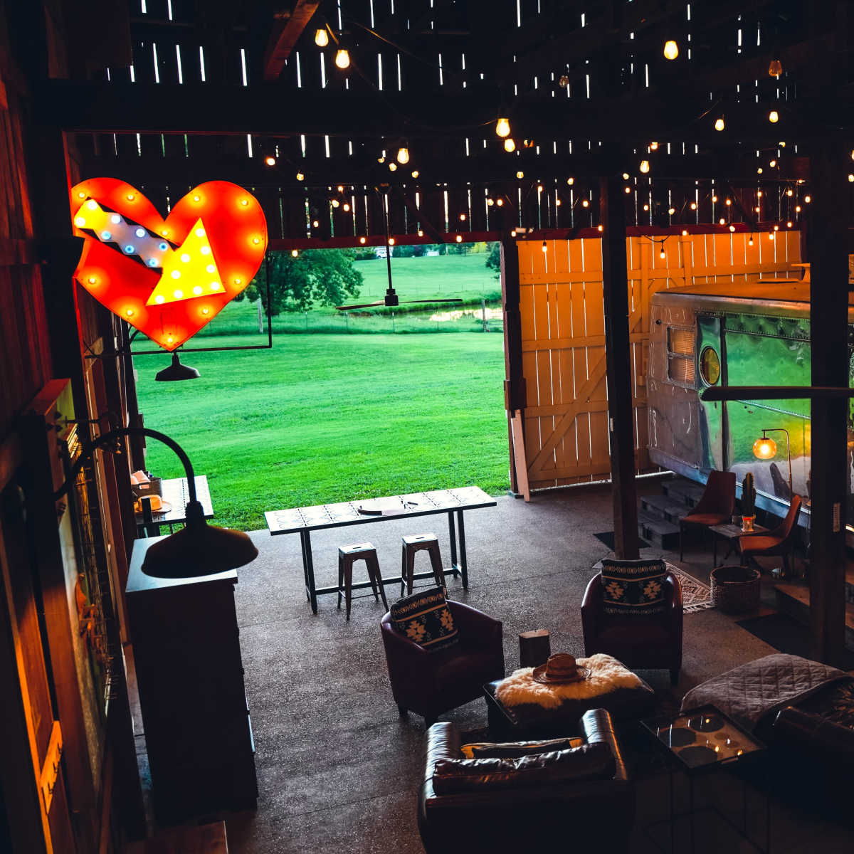 A cozy barn interior with a vintage Airstream trailer, a table, and seating arrangements, illuminated by string lights and a large heart-shaped sign.