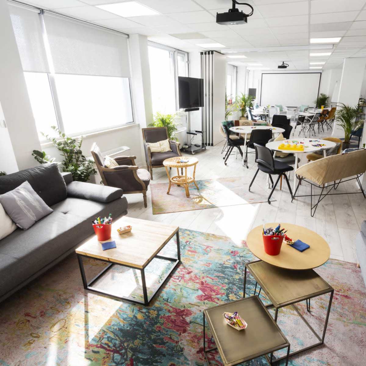 A bright and modern workspace features comfortable seating areas, tables for meetings, and plants throughout the room.