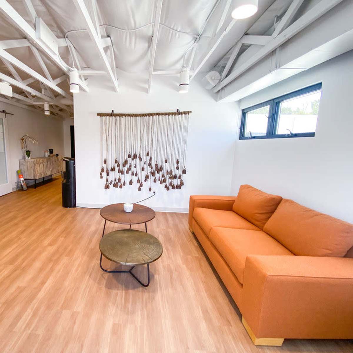 A modern interior space featuring an orange couch, two round tables, and a decorative wall hanging, with wooden flooring and exposed beams in the ceiling.