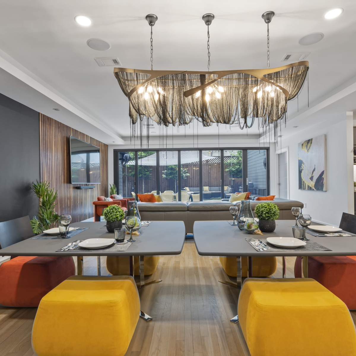 A contemporary dining area features two modern tables with colorful seating, a unique light fixture, and large windows overlooking an outdoor space.