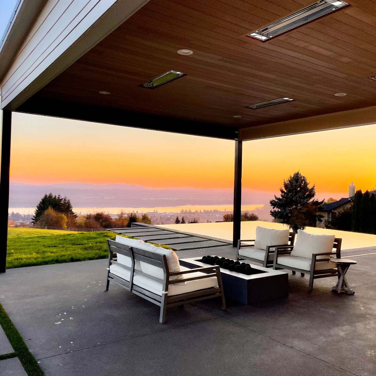 A covered patio with white furniture and a fire pit overlooks a sunset view of a lake and distant hills.