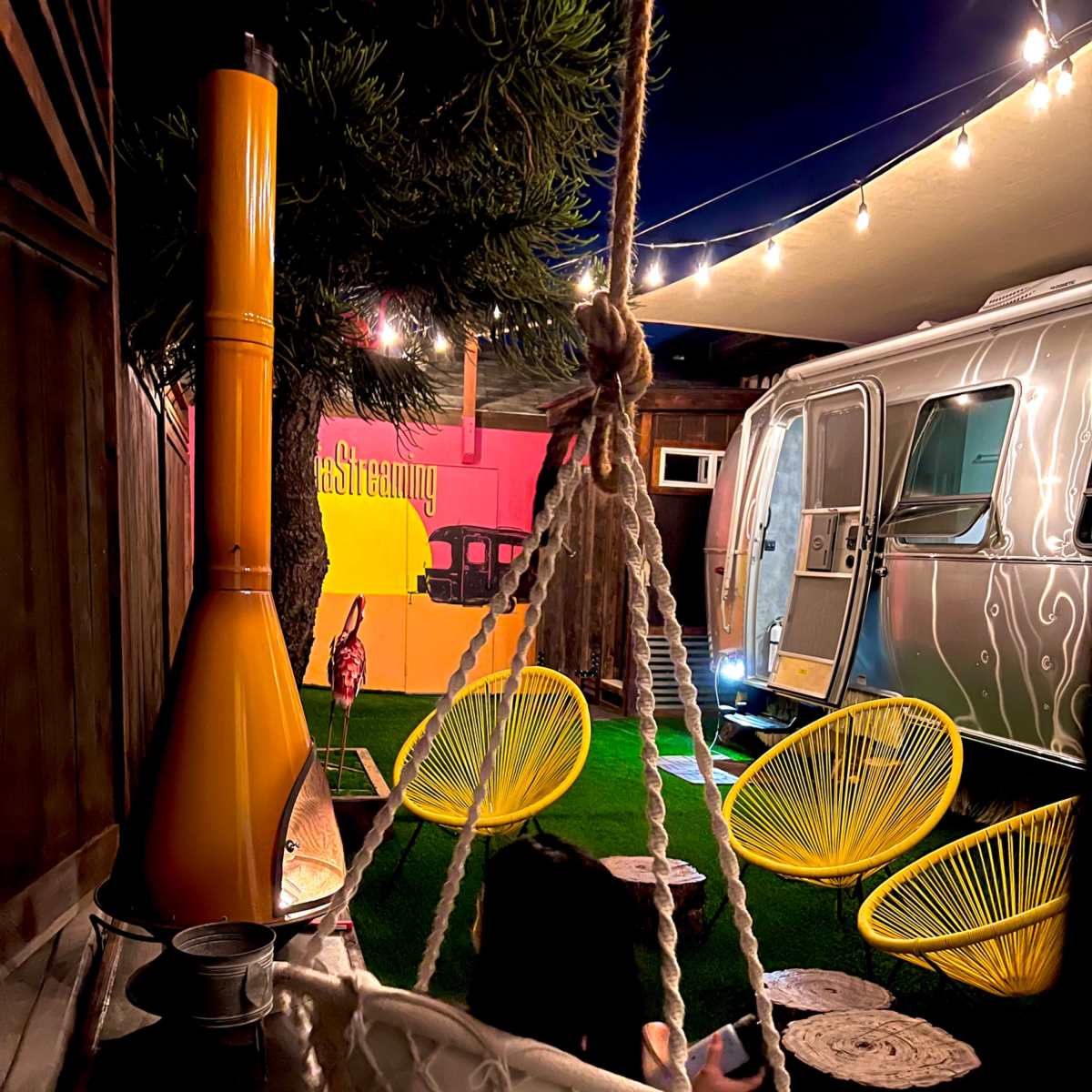 A person sits in a white hanging chair surrounded by bright green grass, colorful decorations, and an Airstream trailer under string lights at night.