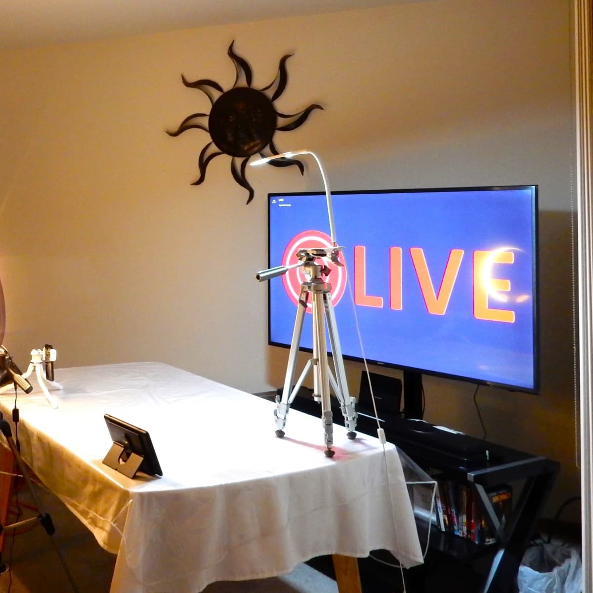 The scene shows a setup for a live broadcast, featuring a table covered with a white cloth, a television displaying "LIVE," and a backdrop of dark curtains.