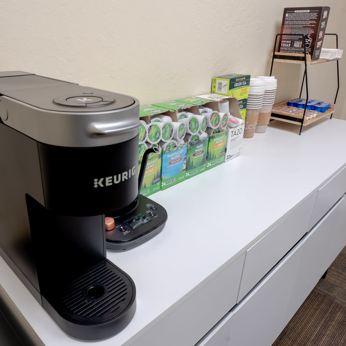 A Keurig coffee maker on a white counter with a selection of coffee pods, cups, and condiments arranged nearby.