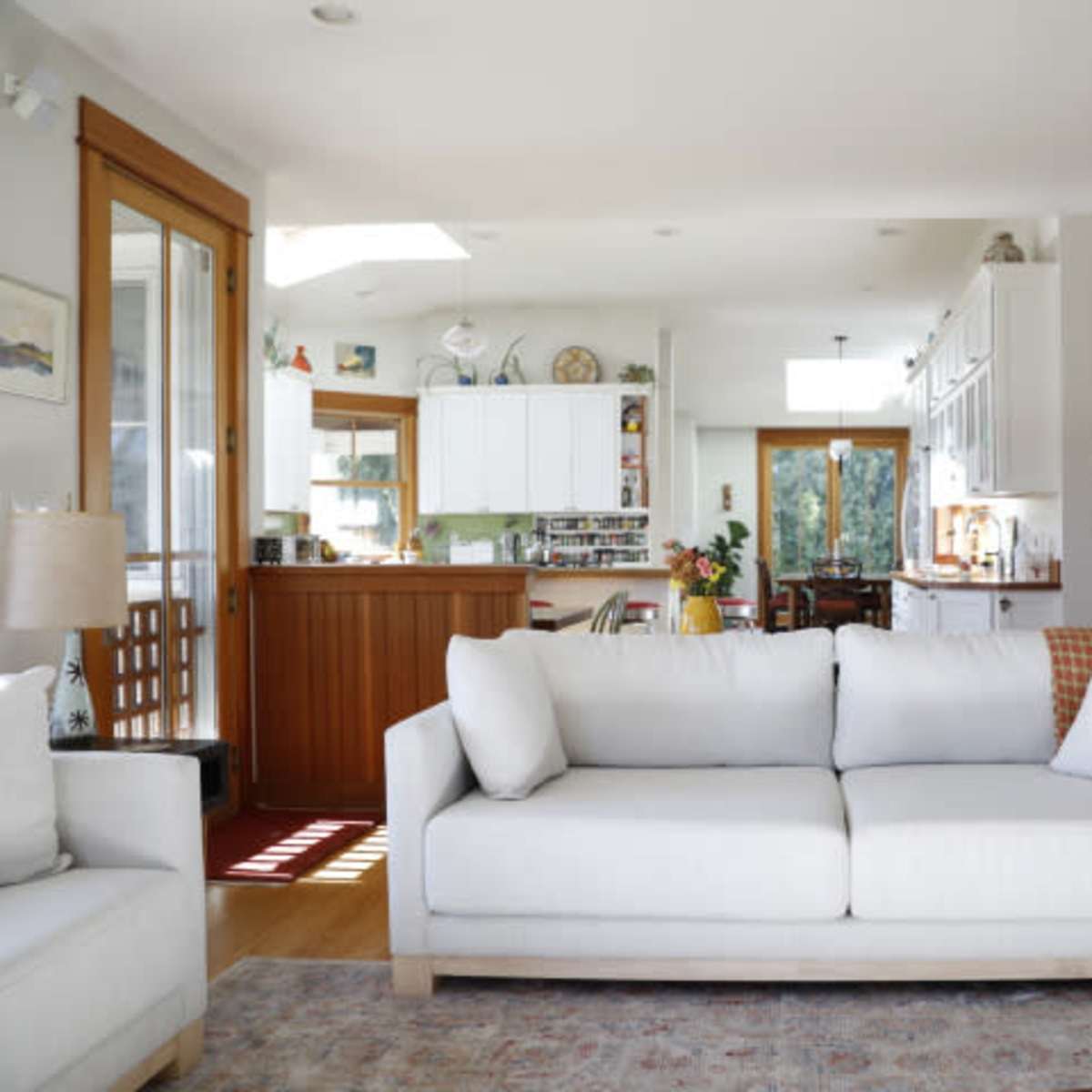 A light-filled living room features a white sofa facing a kitchen with wooden cabinetry and a staircase in the background.