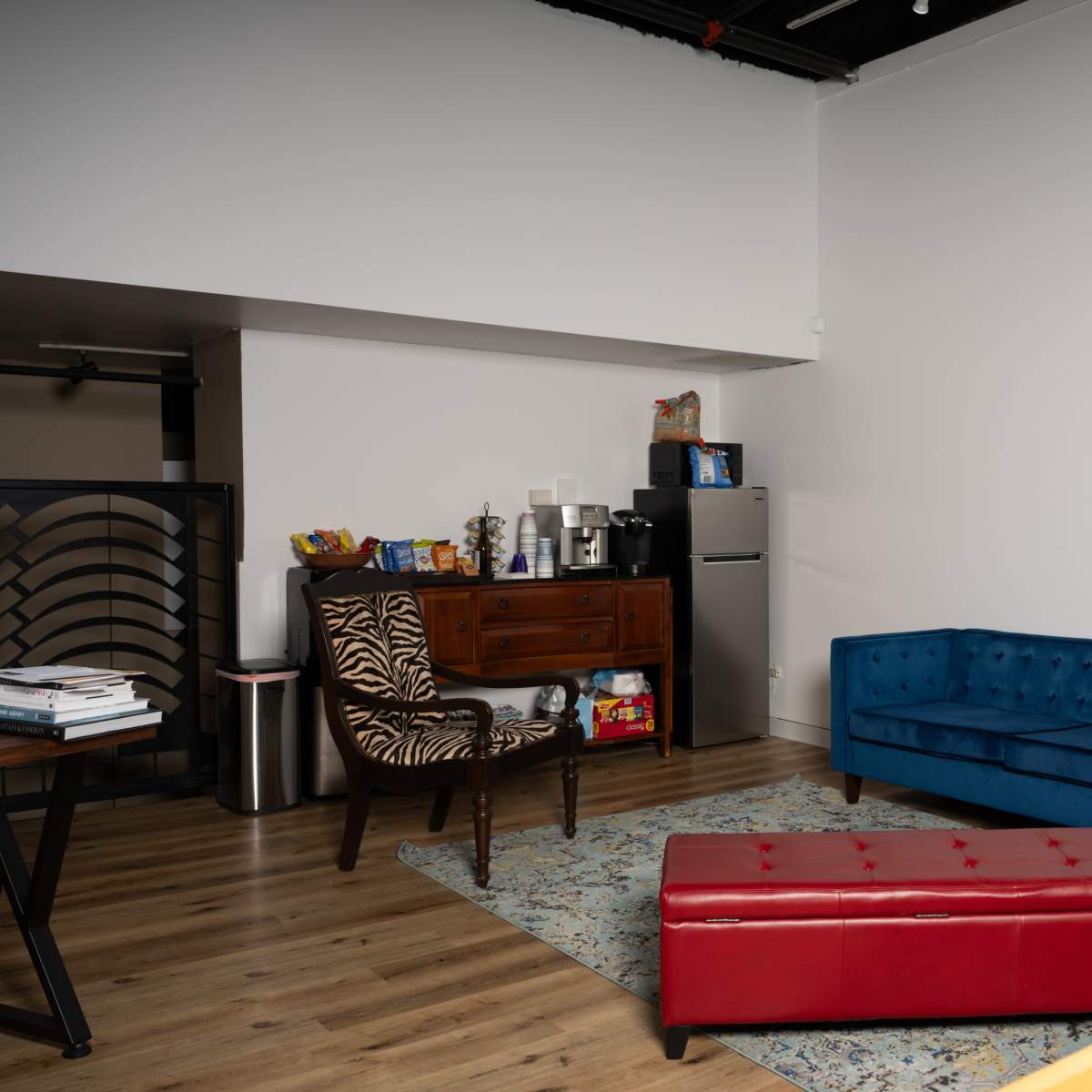 A modern, minimalistic living space featuring a blue sofa, a red ottoman, a zebra-patterned chair, and a rustic wooden cabinet.