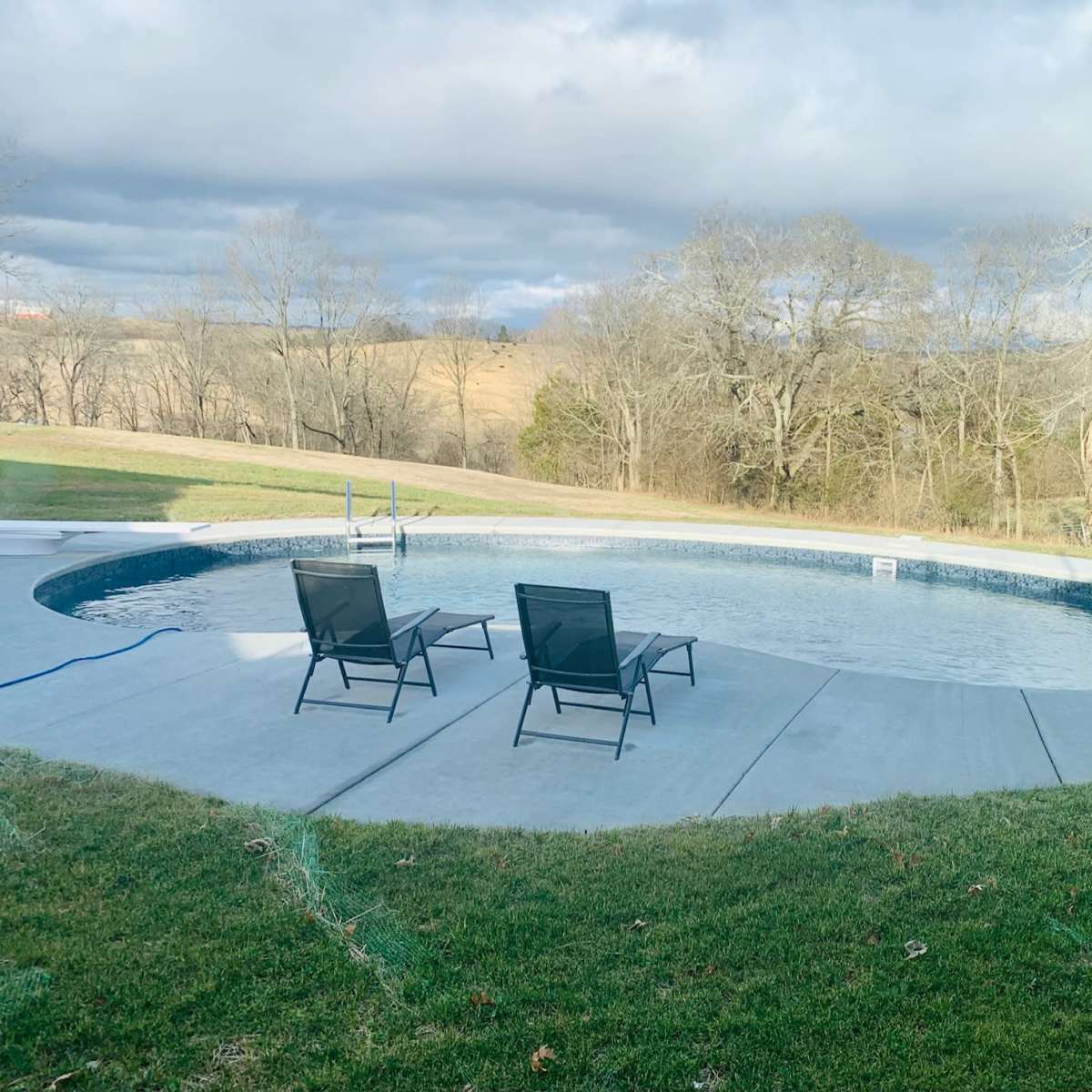 A swimming pool surrounded by a concrete deck and two black lounge chairs, with bare trees and rolling hills in the background under a cloudy sky.