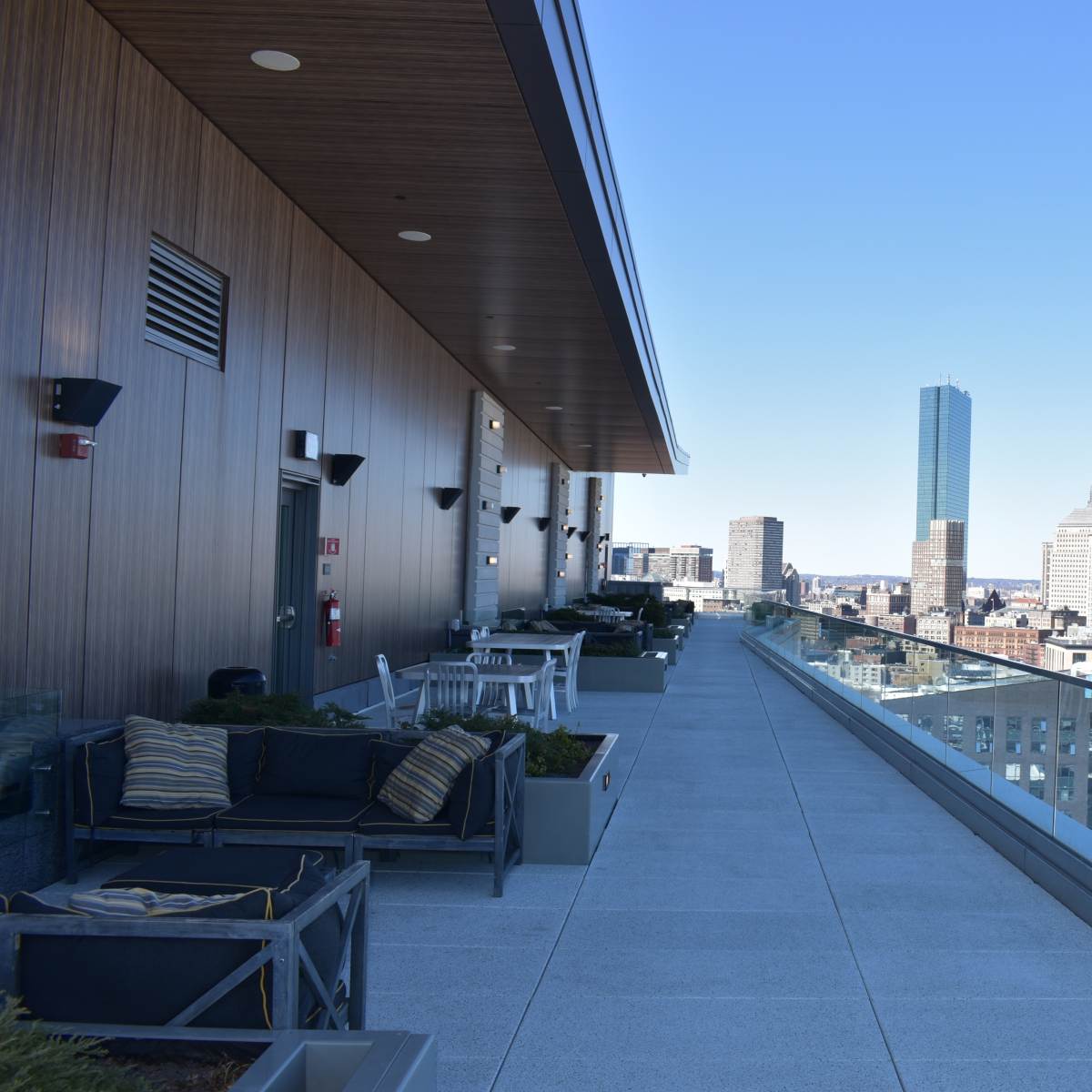A spacious rooftop terrace with modern furniture, overlooking a city skyline featuring tall buildings and a clear blue sky.