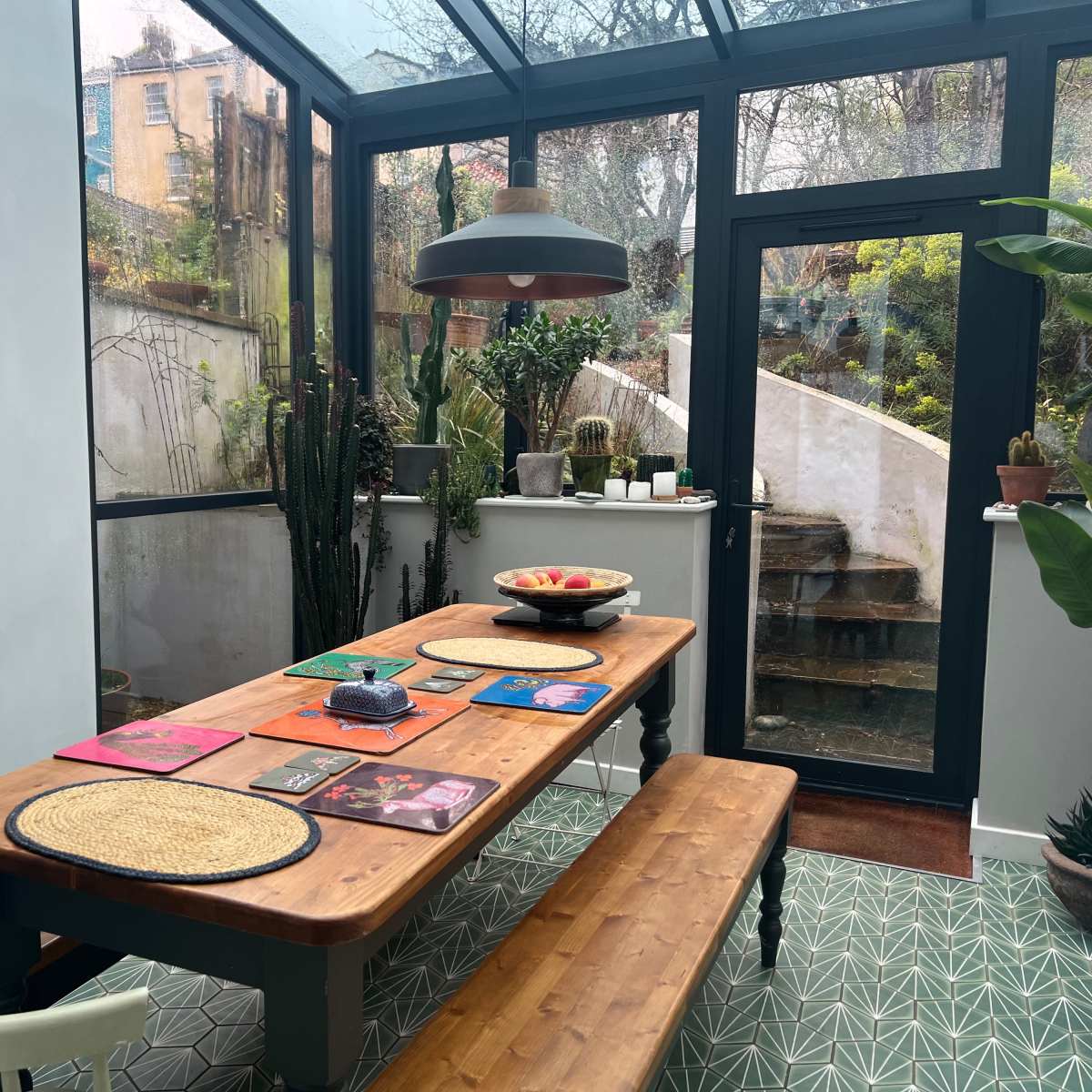 A wooden dining table with benches is set in a bright room featuring large glass windows and a view of a garden, adorned with plants and artwork on the walls.