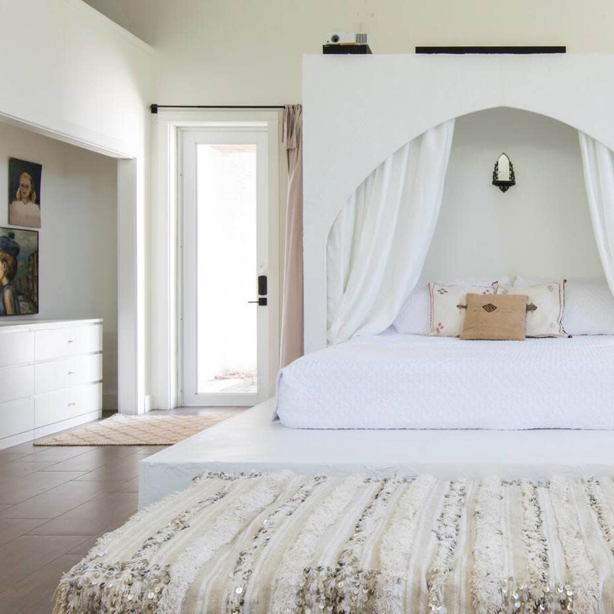 A modern bedroom with a raised, white four-poster bed draped in curtains, positioned on a light-colored floor with a decorative rug in front.