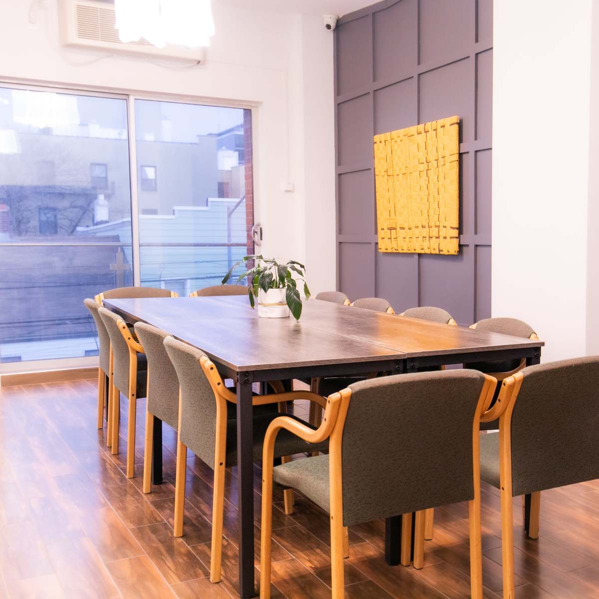 A large conference room features a long wooden table surrounded by ten chairs, with a plant on the table and a large window overlooking an exterior balcony.