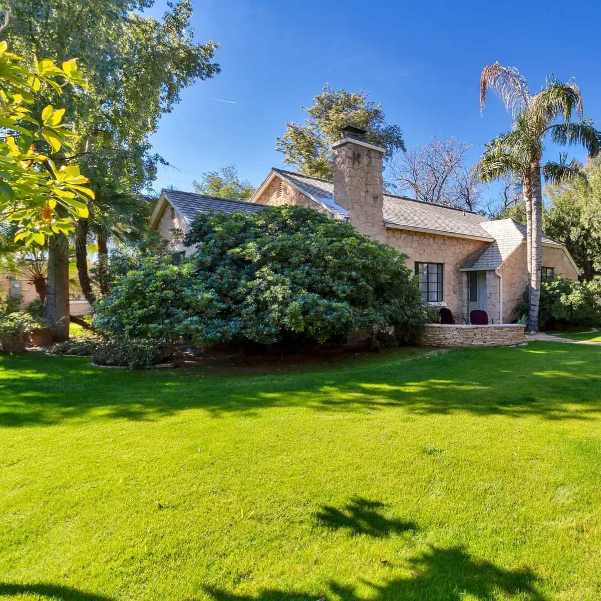 A small house with a stone chimney is surrounded by a well-maintained lawn and several trees.
