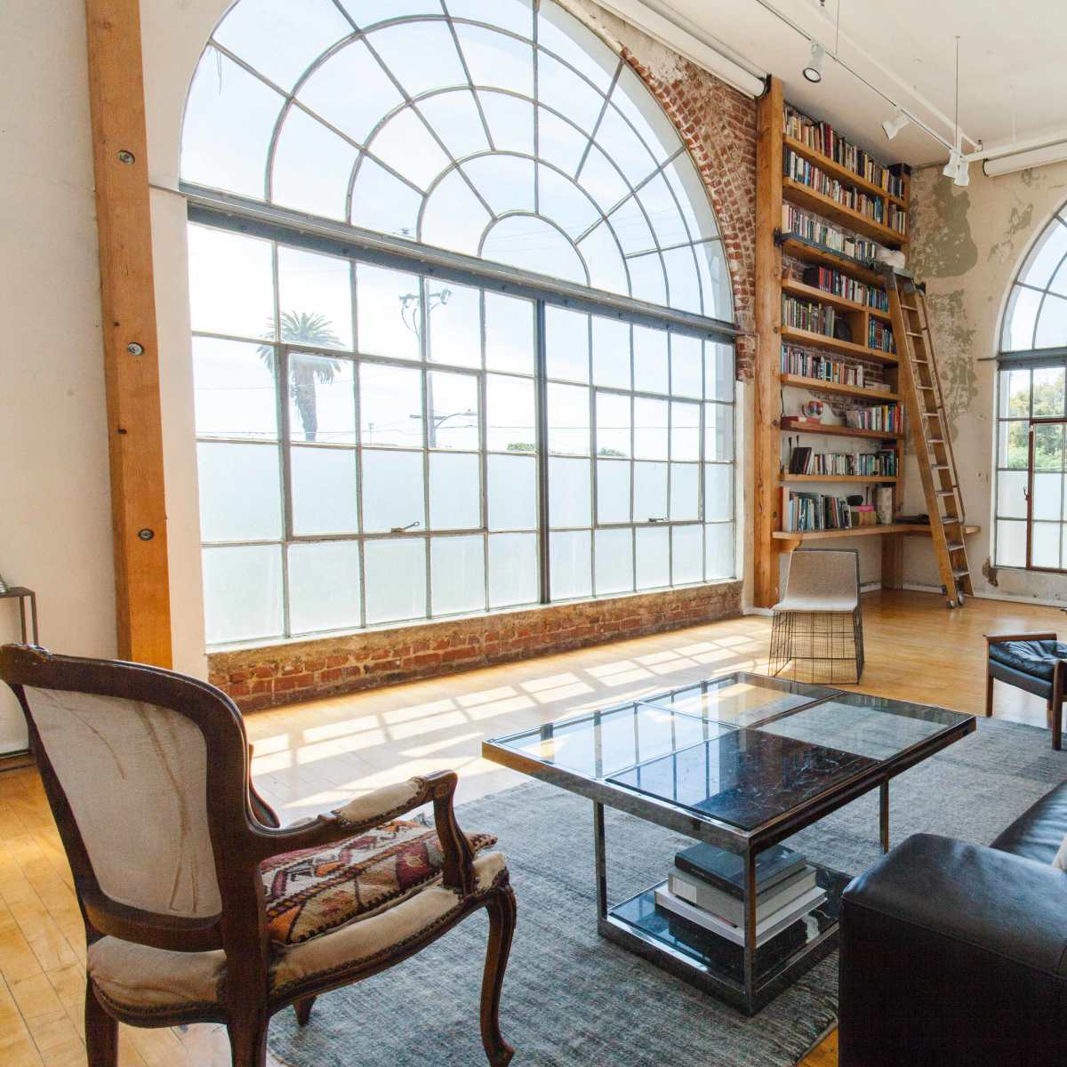 A spacious room with large arched windows, a bookshelf on a ladder, a coffee table, and a seating area with black leather chairs and a patterned armchair.