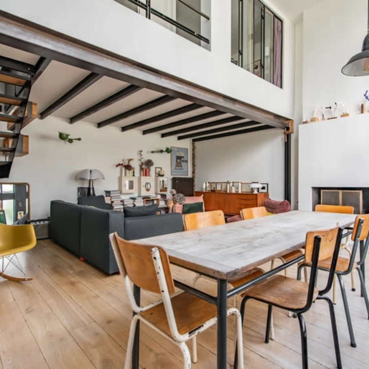 A spacious living area featuring a large wooden dining table surrounded by metal and wooden chairs, with a seating area visible in the background and a staircase leading to an upper level.