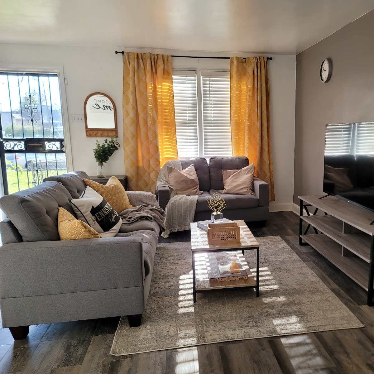 A living room with a gray couch, decorative pillows, a coffee table, a television on a stand, and large windows covered by yellow curtains.