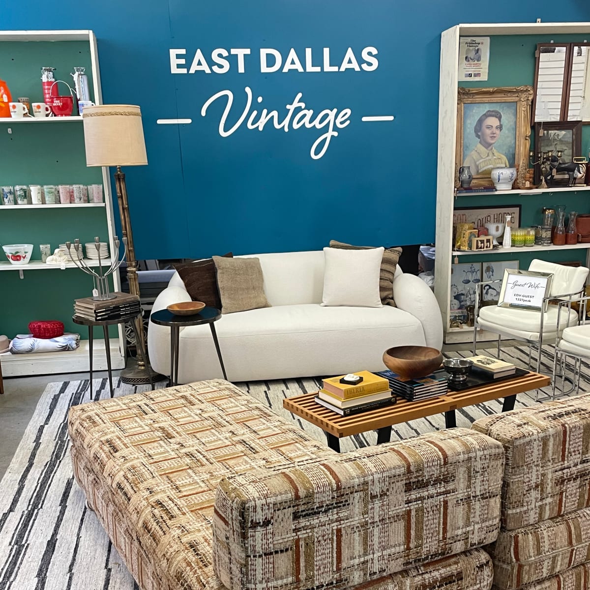A vintage furniture display featuring a white couch, a patterned sectional sofa, and various decorative items against a teal wall with "EAST DALLAS Vintage" written on it.