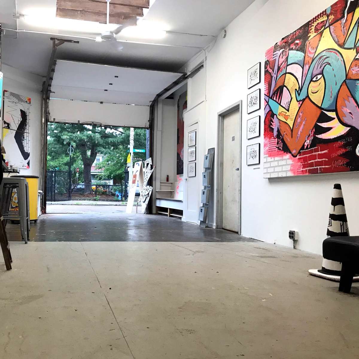 An industrial-style gallery space with an open garage door leading to a green outdoor area, featuring artworks on the walls and various items arranged inside.