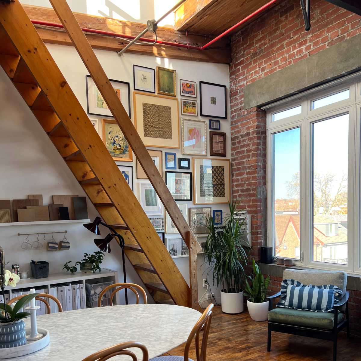 A bright room features a wooden staircase, a round marble table with four chairs, and a wall decorated with various framed artworks.