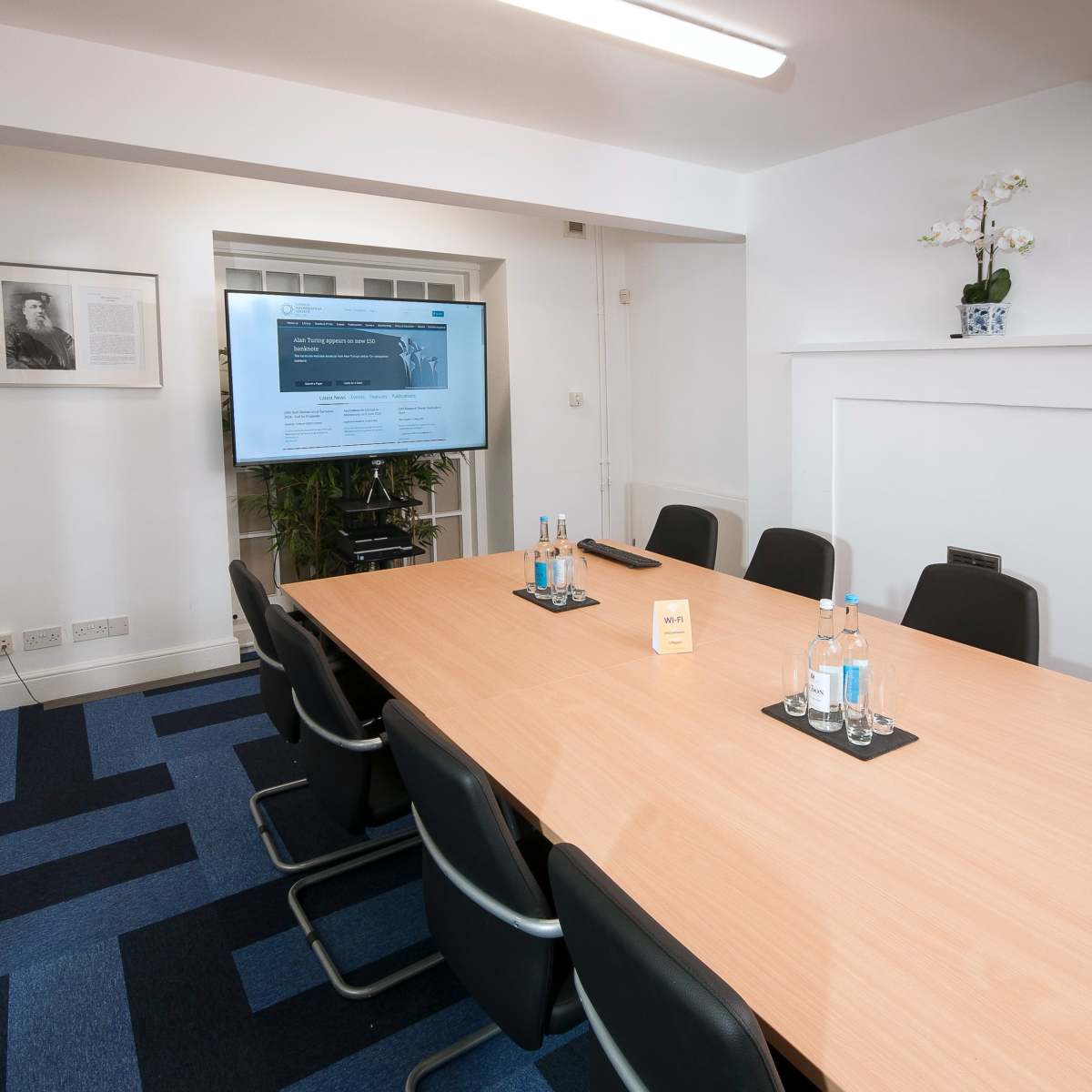 A modern conference room features a long wooden table surrounded by black chairs, with a large screen displaying a presentation and bottled water on the table.