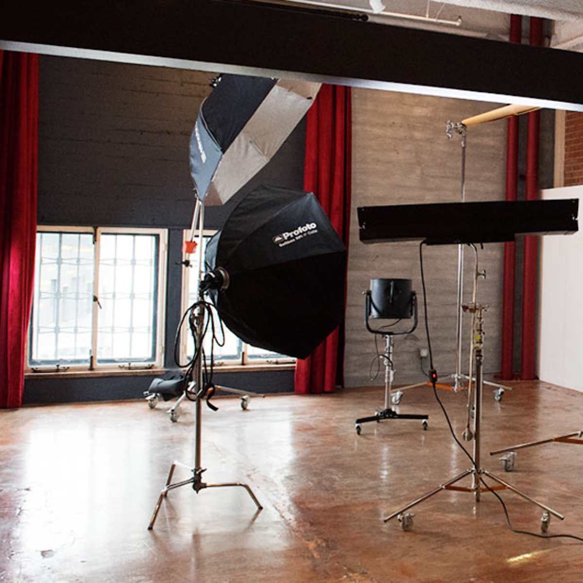 A photography studio with umbrellas, softboxes, and a backdrop setup, featuring a chair and large windows with red curtains.