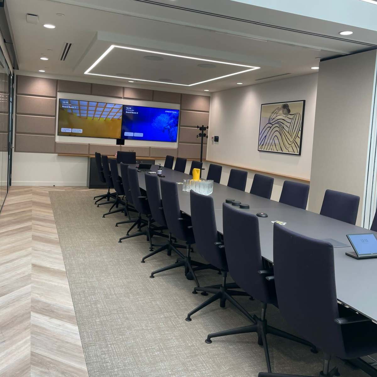A modern conference room with a long table, black chairs, and two screens mounted on the wall.