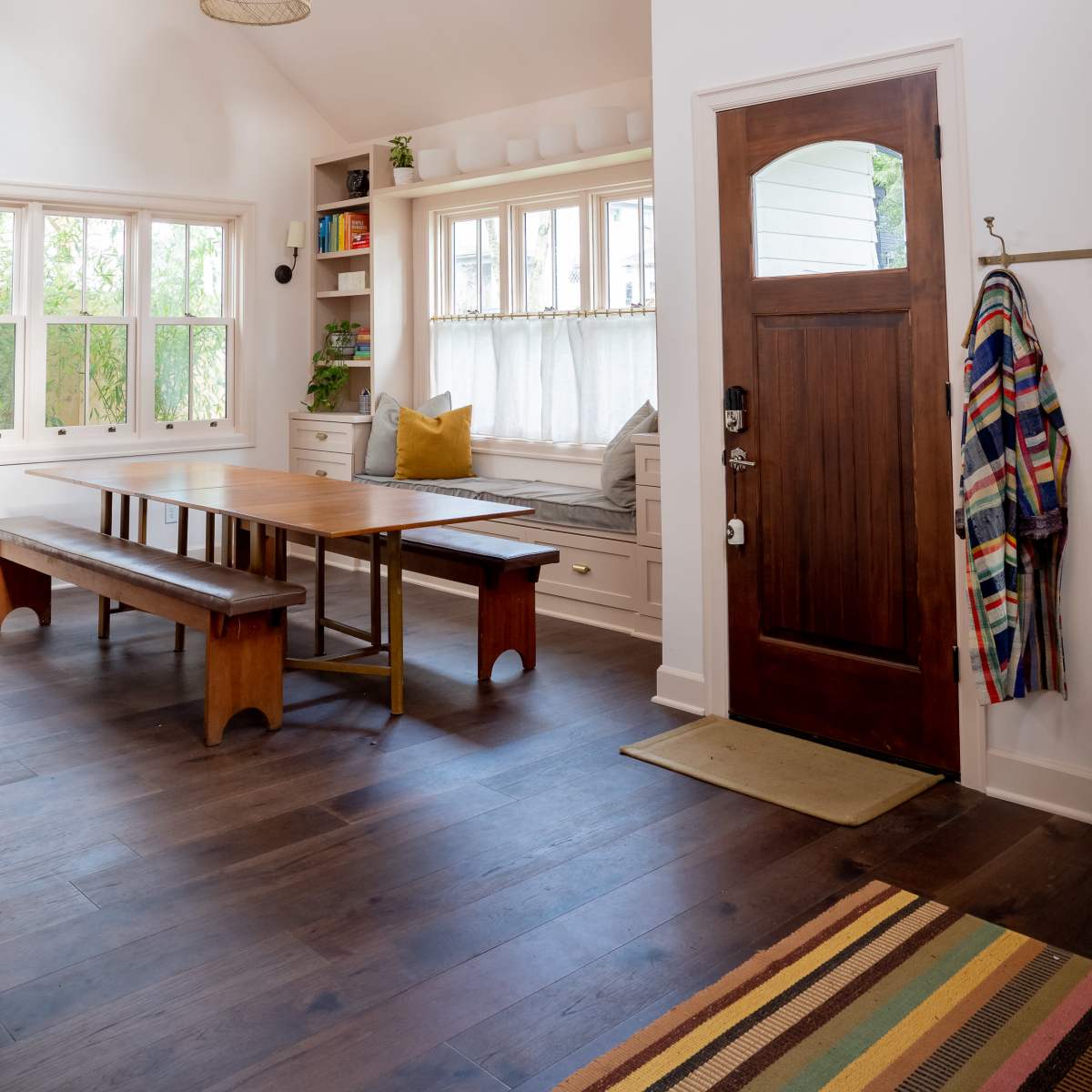 A bright, modern entryway with a wooden front door, a dining table, and a cozy reading nook by the windows.