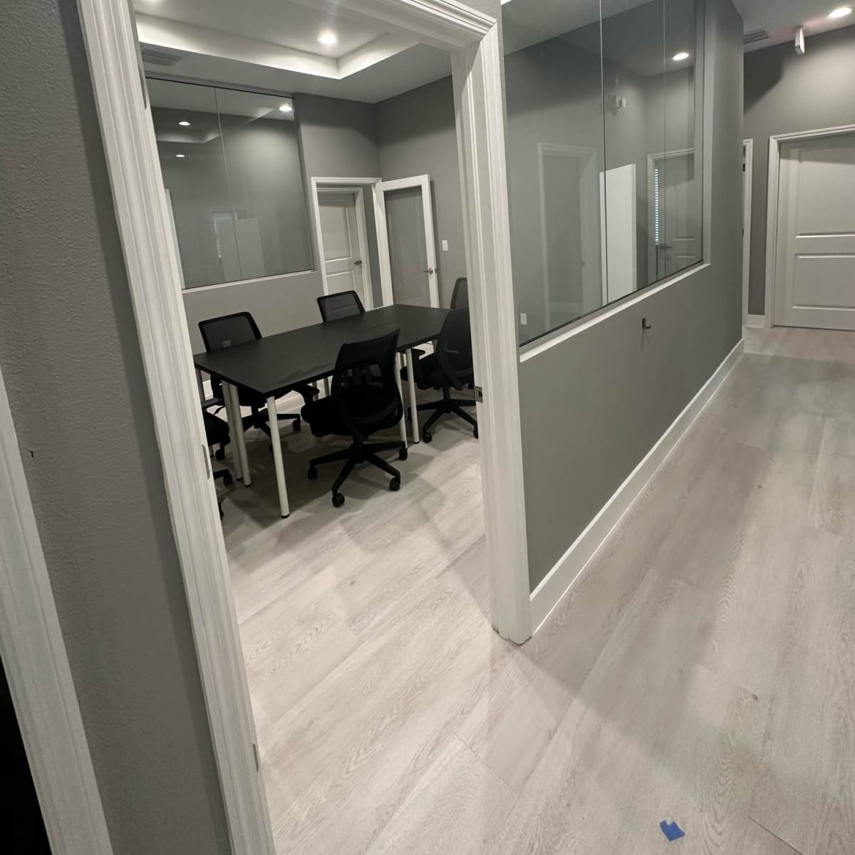 A modern office corridor leading to a conference room with a large table and several chairs, visible through a glass wall.