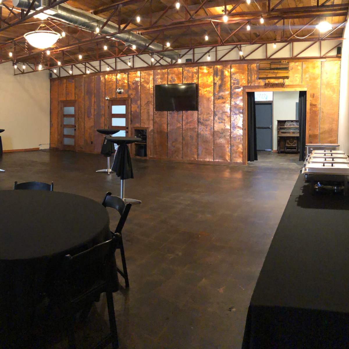 A spacious event room with dark flooring, a copper accent wall, and several tables and stands set up for an event.