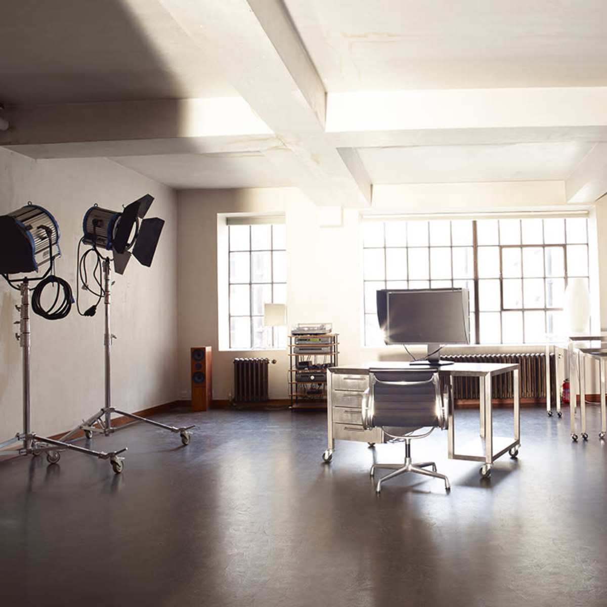 A spacious, minimalist studio with a desk, a computer monitor, and multiple lighting setups beside large windows.