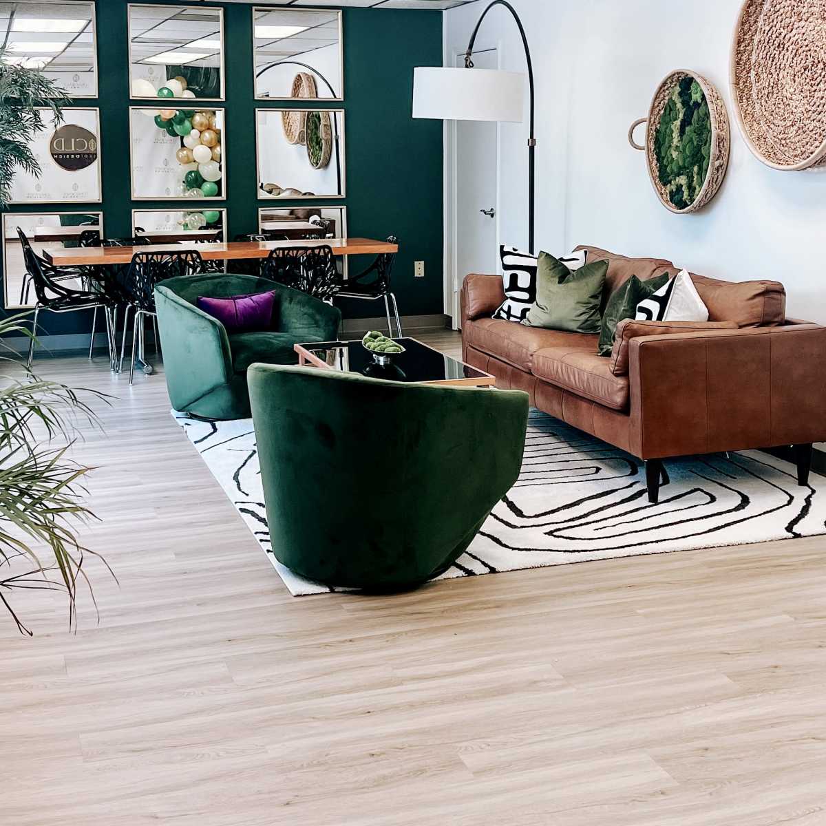 A modern office lounge featuring a brown leather sofa, green armchairs, a patterned rug, and decorative plants, arranged around a central area with a dining table and chairs.