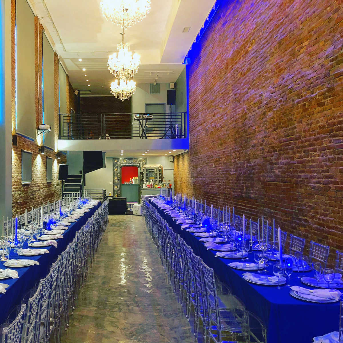 A long dining table set for an event in a brick-walled hall, illuminated by chandeliers and blue lighting.