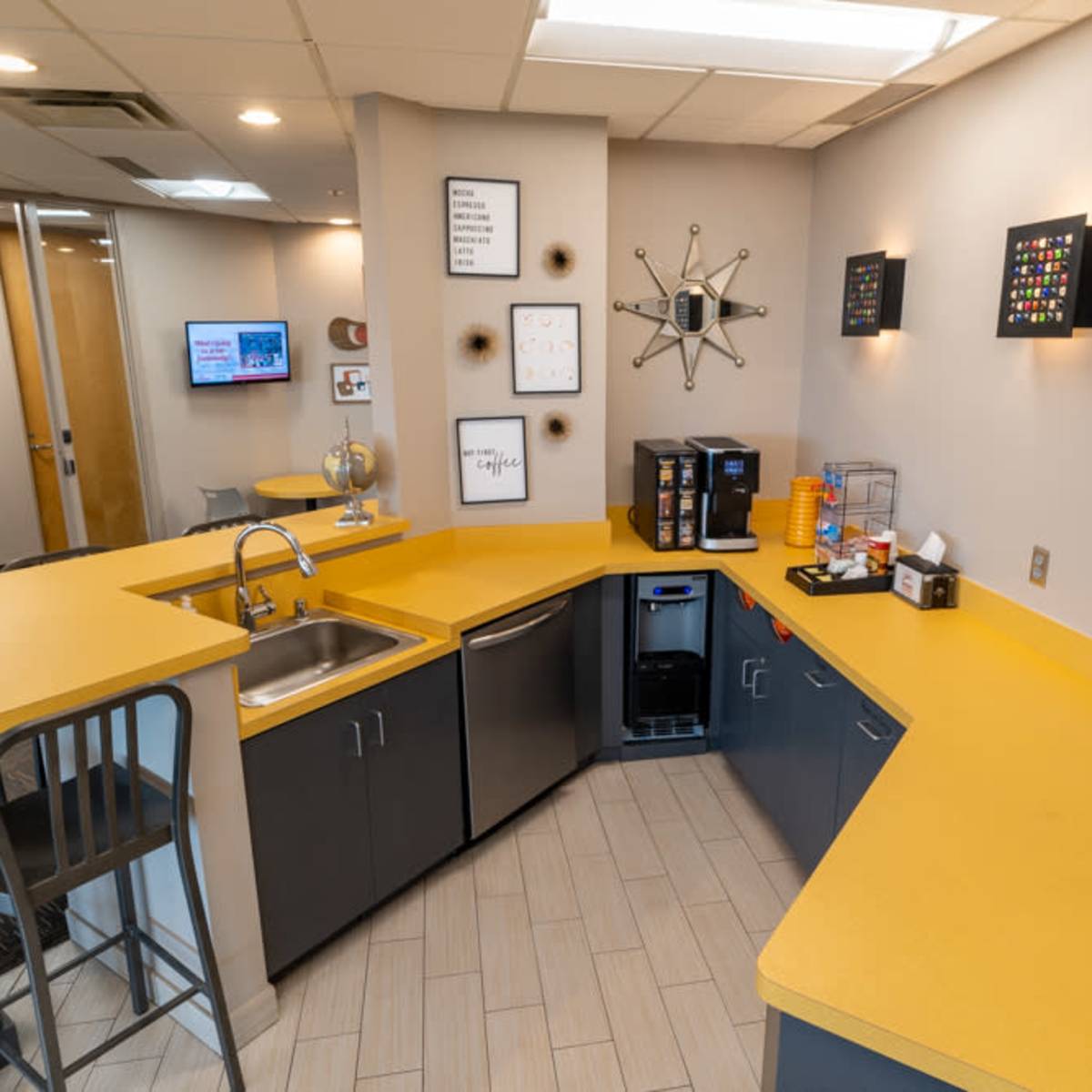 A modern kitchen area with a yellow countertop, a sink, a coffee machine, and various wall decorations.