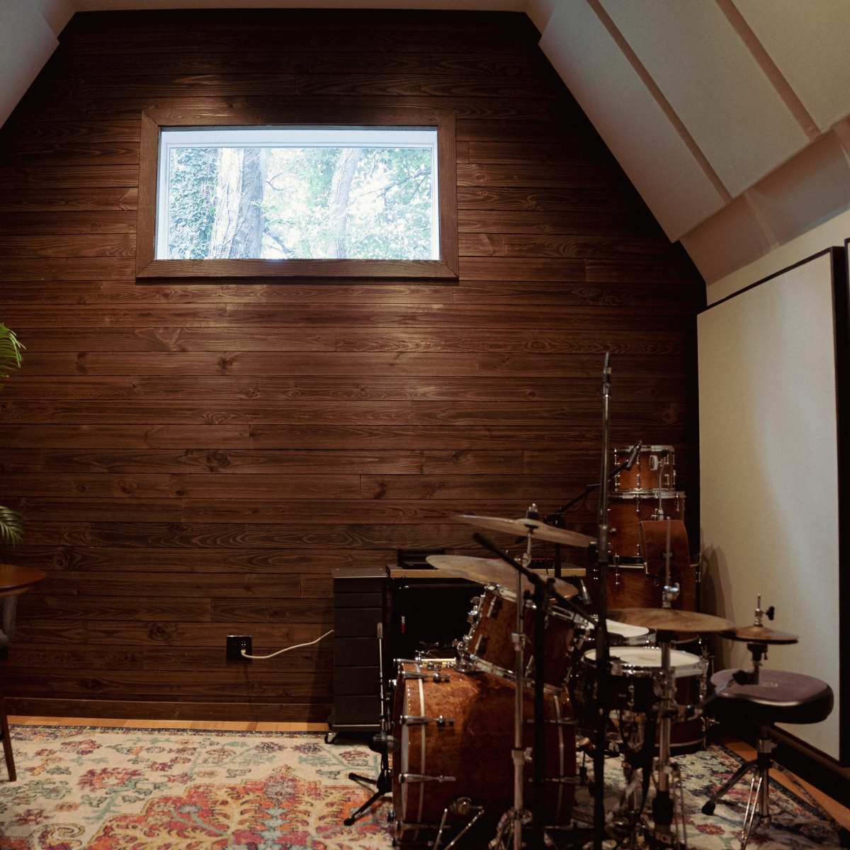 A small music studio featuring a drum set, a table with chairs, and a large window above a wooden wall with a view of trees outside.