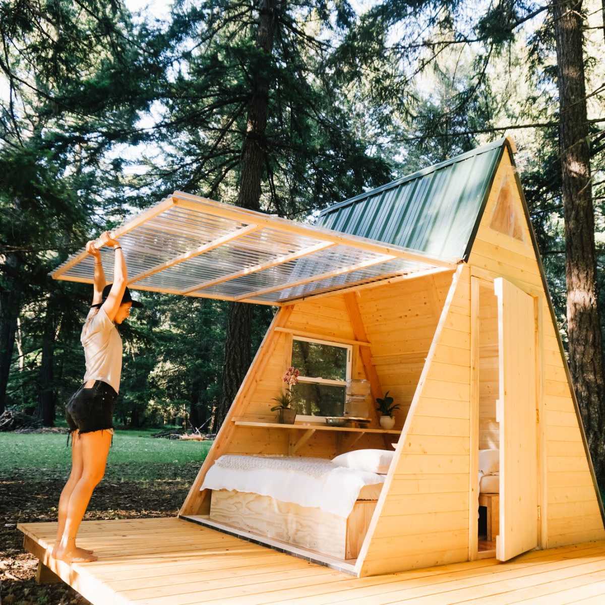 A person raises the roof of a small wooden A-frame cabin in a forest clearing.