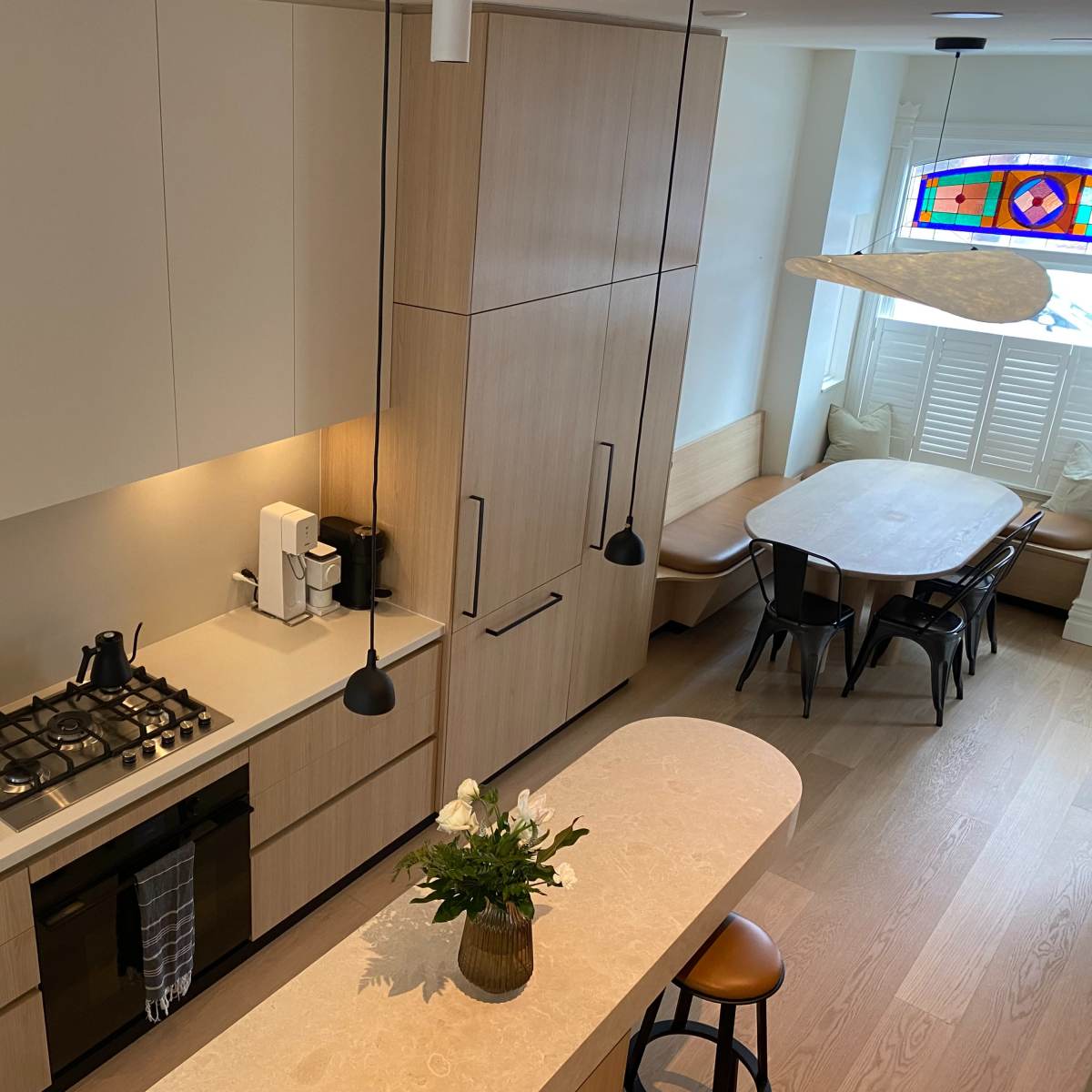 A modern kitchen with wooden cabinets, a gas stove, and a dining area featuring a large table and a colorful stained glass window.