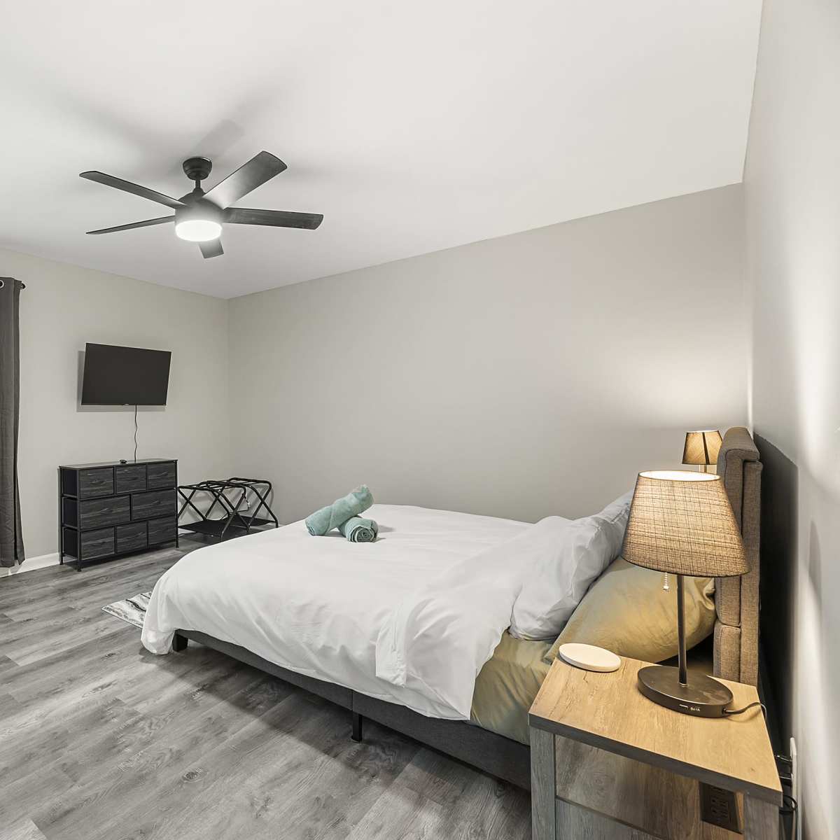A well-lit bedroom featuring a bed with white bedding, a nightstand with a lamp, a dresser, a TV mounted on the wall, and large windows with blinds.